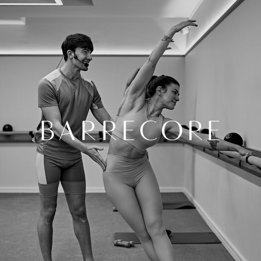 Barrecore instructor providing hands-on alignment cues during a barre class, supporting precise posture and controlled movement at the barre in a results-driven, low-impact workout.