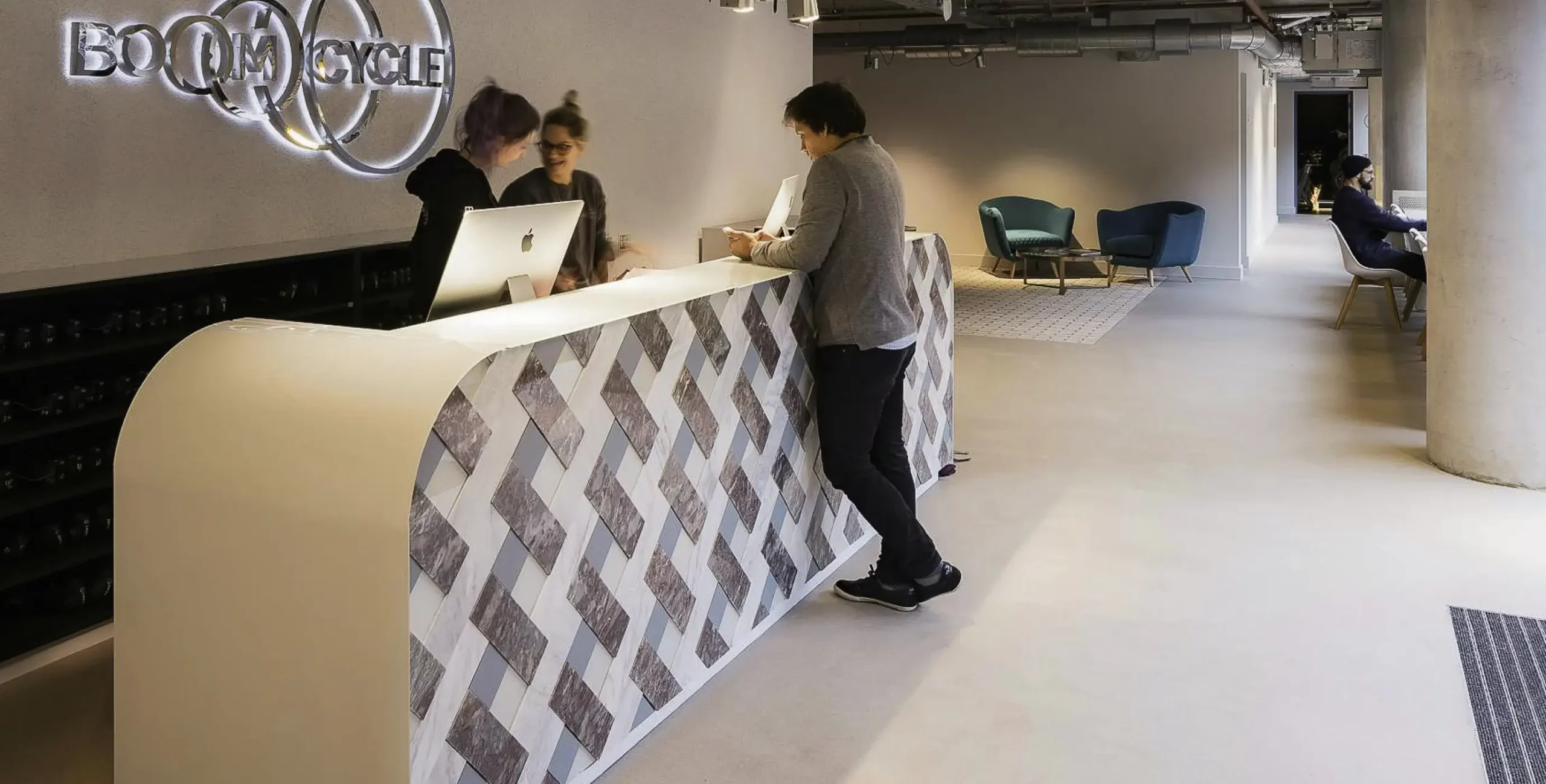 Reception area at Boom Cycle studio, with staff assisting guests at a modern front desk under a neon logo, and lounge seating visible in the background.