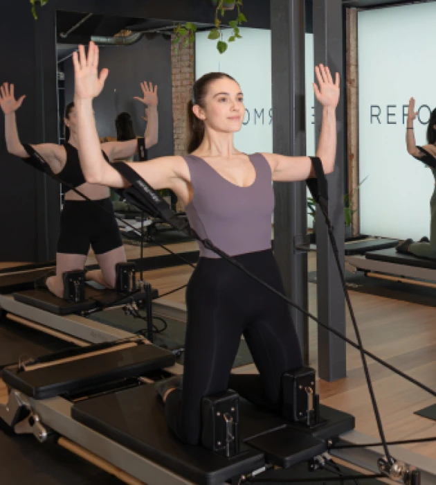 Client performing kneeling arm work with resistance straps on the Reformer at Reformcore, targeting posture, shoulder strength, and full-body alignment in a modern Pilates studio.