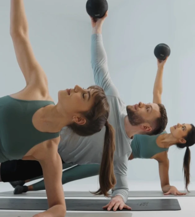 Barrecore clients in a signature class holding side planks with weighted balls, combining isometric strength, control, and precision in a refined, low-impact barre workout.